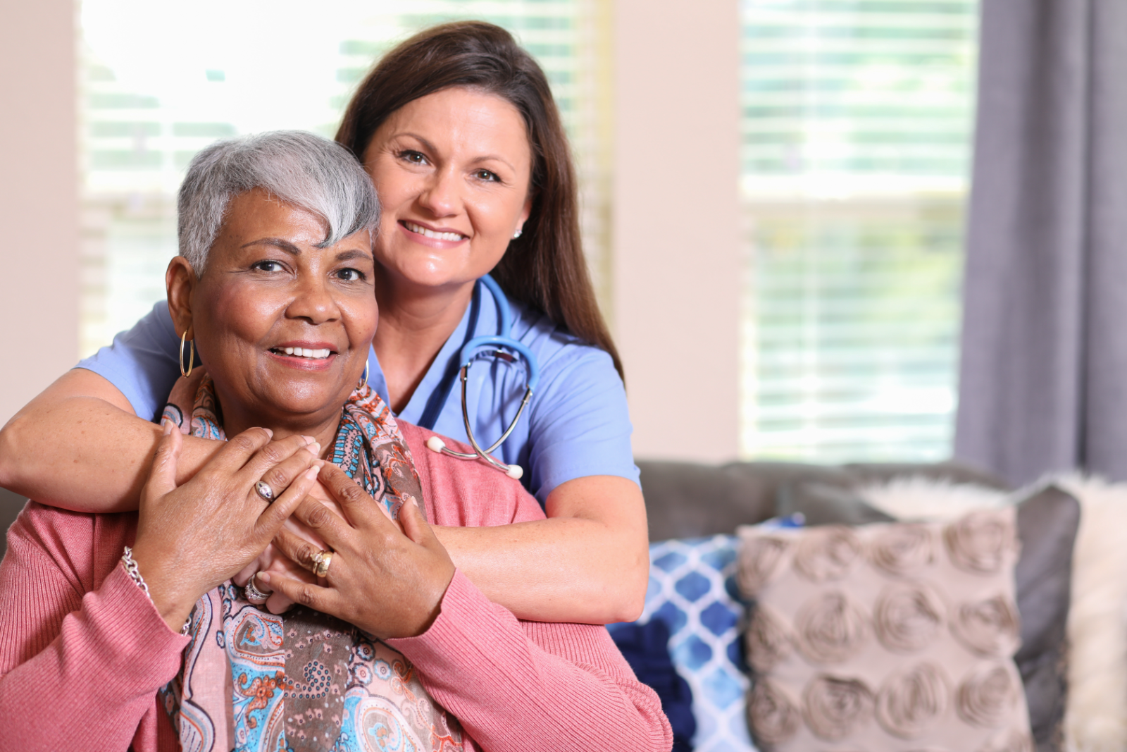 Nurse with her arms around a smiling older woman on her living room sofa